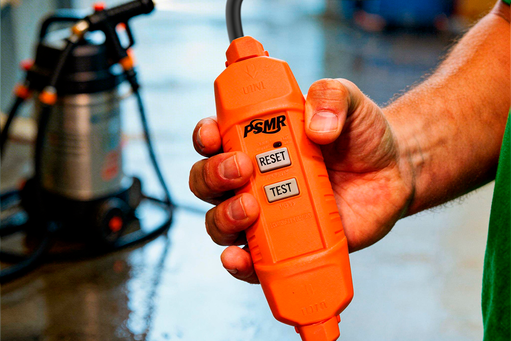 Mão segurando IDR móvel PSMR laranja com botões RESET e TEST, em ambiente molhado com lavadora de alta pressão ao fundo